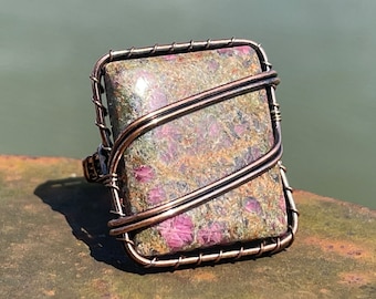 May include: A handcrafted ring featuring a rectangular stone with a speckled pattern of green, brown, and pink hues. The stone is encased in a copper-colored wire frame, creating a unique and artistic design. The ring is a statement piece.