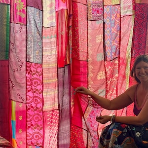 May include: A patchwork curtain made of various pink and red fabrics. The curtain is composed of rectangular panels with different patterns and textures. The fabrics include floral designs and geometric shapes. The curtain is hanging from a rod.