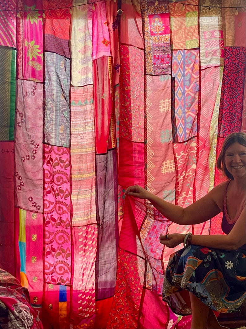 May include: A patchwork curtain made of various pink and red fabrics. The curtain is composed of rectangular panels with different patterns and textures. The fabrics include floral designs and geometric shapes. The curtain is hanging from a rod.