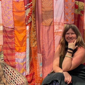 以下が含まれることがあります： カラフルなパッチワークのカーテンの前で、籐の椅子に座っている女性。カーテンはオレンジ、赤、ピンク、紫の様々な色合いで作られています。女性は花柄の黒いドレスと緑色のスカーフを身につけています。