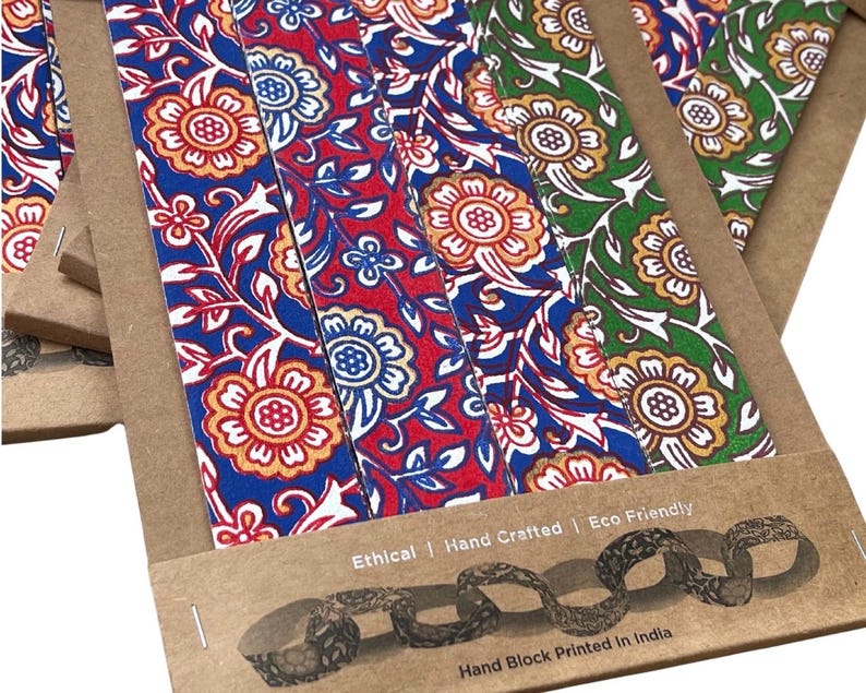 May include: Close-up of several floral patterned bookmarks in blue, red, and green. The bookmarks are made of fabric and are packaged in brown cardboard. The text "Ethical | Hand Crafted | Eco Friendly" and "Hand Block Printed In India" are visible.