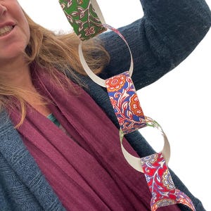May include: A paper chain with alternating links of white and patterned paper. The patterned links feature floral designs in red, blue, and green. The chain is held up against a person wearing a blue sweater and a burgundy scarf.