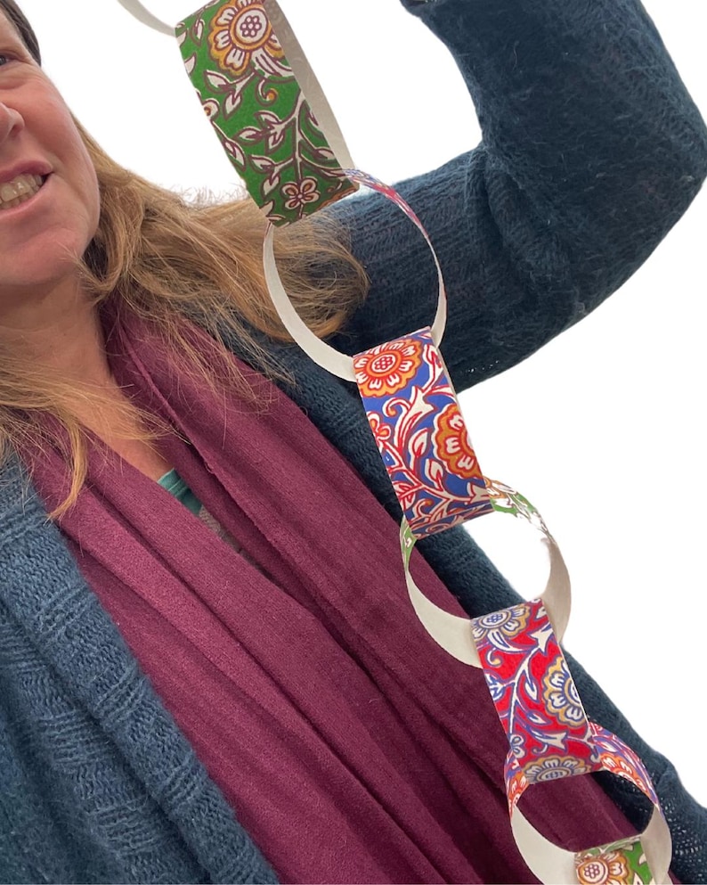 May include: A paper chain with alternating links of white and patterned paper. The patterned links feature floral designs in red, blue, and green. The chain is held up against a person wearing a blue sweater and a burgundy scarf.