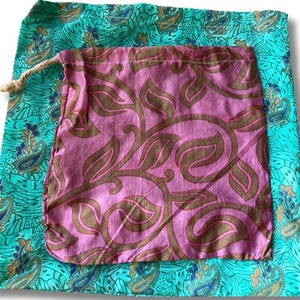 Può includere: Un sacchetto rosa con coulisse e motivo a foglie marroni è appoggiato su un tessuto turchese con un disegno a paisley. La borsa è realizzata in un materiale morbido e ha un cordoncino in corda naturale.
