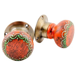May include: Two decorative door knobs. The knobs are painted orange with green and white decorative patterns. The base is a bronze colour. The knobs are designed to be installed on a door.
