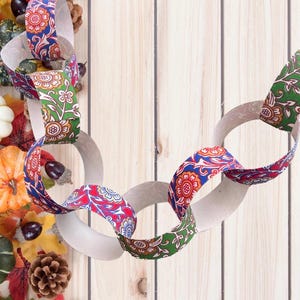May include: A decorative paper chain with links of white paper and patterned fabric. The fabric has a floral design in red, blue, and green. The chain is surrounded by pumpkins, gourds, leaves, and pinecones on a wooden background.