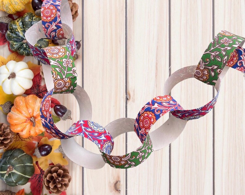 May include: A decorative paper chain with links of white paper and patterned fabric. The fabric has a floral design in red, blue, and green. The chain is surrounded by pumpkins, gourds, leaves, and pinecones on a wooden background.