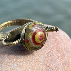 May include: A brass ring with a circular, eye-like design. The central eye is yellow with red and white accents. The ring is set on a light brown stone, with a blurred green and blue background.
