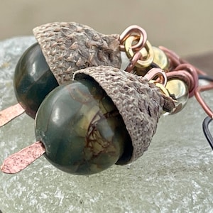 Peut inclure: Gros plan sur des boucles d'oreilles en forme de glands. Chaque boucle d'oreille présente une perle verte foncée et tachetée avec un capuchon marron texturé. Les boucles d'oreilles ont des accents métalliques cuivrés et dorés et des crochets d'oreilles noirs.