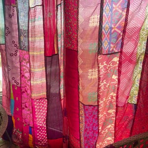 May include: A patchwork curtain made of various fabrics in shades of red, pink, and purple. The fabrics have different patterns, including floral, geometric, and abstract designs. The curtain is hanging, and the light shines through the sheer materials.