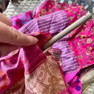 May include: A close-up of a patchwork textile in shades of pink, purple, and gold, with a wooden dowel inserted. The fabric features various patterns and textures, including stripes and floral designs. The edges are stitched with yellow thread.