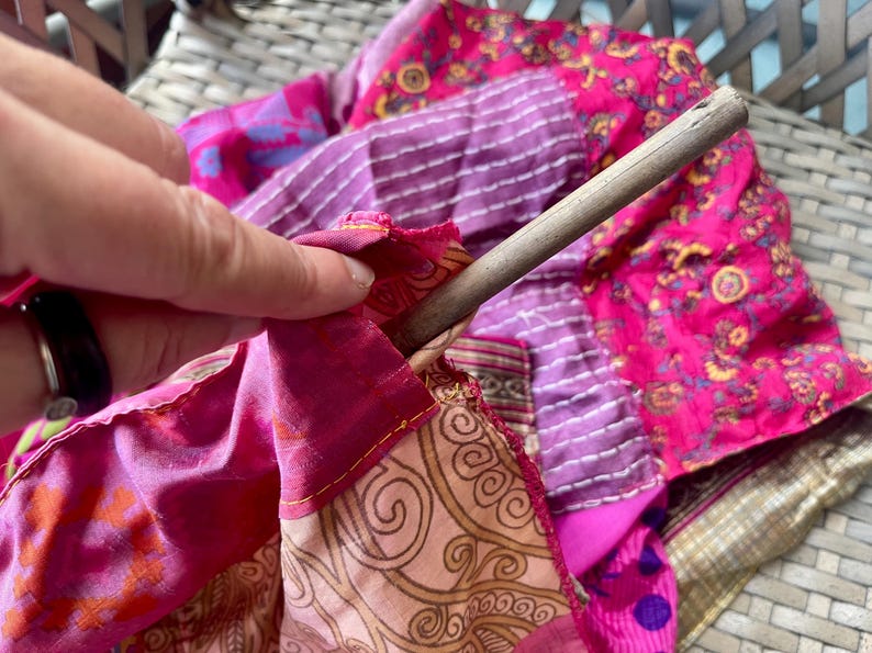 May include: A close-up of a patchwork textile in shades of pink, purple, and gold, with a wooden dowel inserted. The fabric features various patterns and textures, including stripes and floral designs. The edges are stitched with yellow thread.