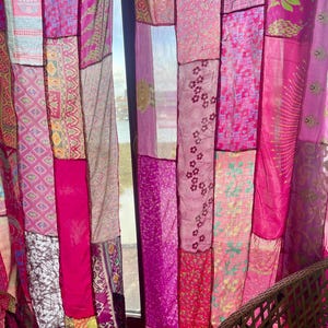 May include: Patchwork curtain panels in shades of pink and magenta. The panels are made from various fabrics with floral and geometric patterns. The curtain hangs in front of a window, with a glimpse of the outside.