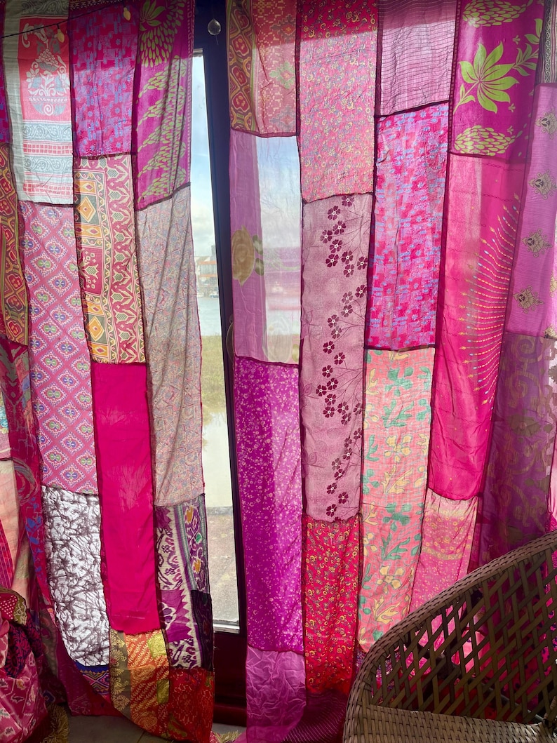 May include: Patchwork curtain panels in shades of pink and magenta. The panels are made from various fabrics with floral and geometric patterns. The curtain hangs in front of a window, with a glimpse of the outside.