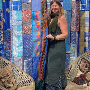 Peut inclure: Une femme portant une robe vert foncé avec une jupe à imprimé floral se tient devant un rideau de patchwork coloré. Le rideau est fait de différents tissus dans des tons de bleu, orange et rose. La femme sourit et a de longs cheveux bruns.