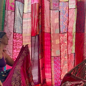 May include: A patchwork curtain made of various fabrics in shades of pink and red. The curtain is hanging, with different patterns and textures visible. The fabrics appear to be lightweight, possibly silk or a similar material. The overall aesthetic is bohemian and colorful.