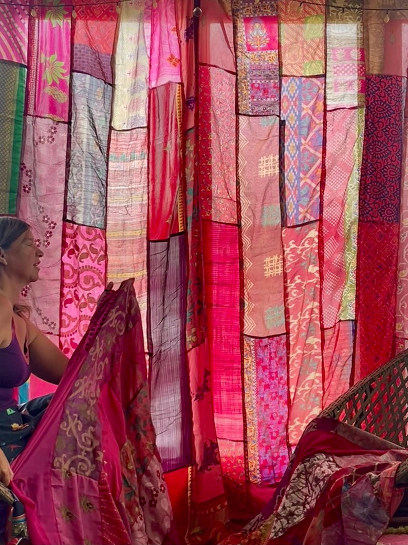 May include: A patchwork curtain made of various fabrics in shades of pink and red. The curtain is hanging, with different patterns and textures visible. The fabrics appear to be lightweight, possibly silk or a similar material. The overall aesthetic is bohemian and colorful.