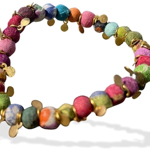 Pode incluir: Um bracelete de tecido colorido com detalhes dourados. O bracelete é feito de pequenas contas redondas que são cobertas de tecido. As contas são enfiadas com fio dourado e têm pingentes dourados que pendem.
