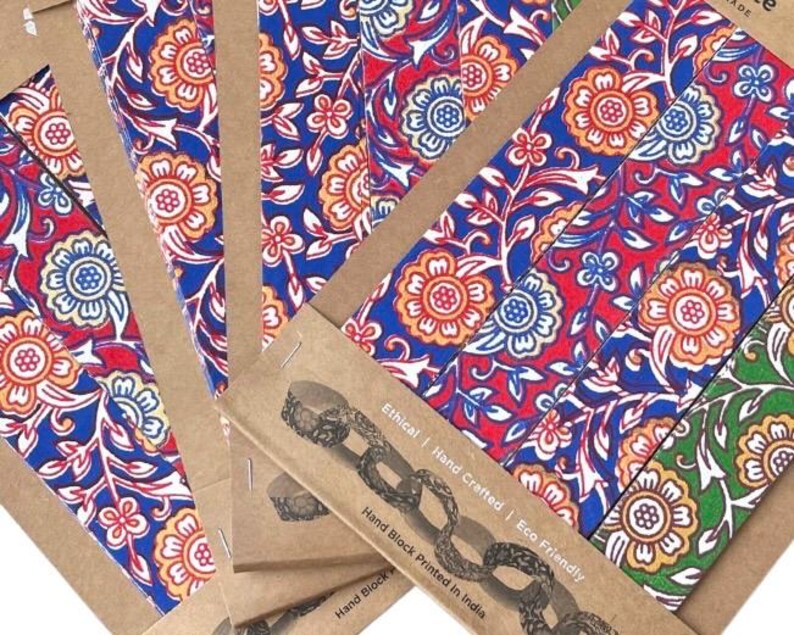 May include: Close-up of several notebooks with colorful floral patterns in red, blue, and orange. The notebooks are in cardboard covers with text that reads "Ethical | Hand Crafted | Eco Friendly" and "Hand Block Printed in India."