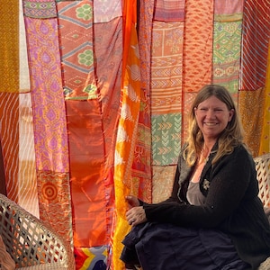 Pode incluir: Uma mulher est&aacute; sentada em uma cadeira de vime em frente a uma cortina de patchwork colorida. A cortina &eacute; feita de muitos tecidos diferentes em tons de laranja, vermelho, amarelo e roxo. A mulher est&aacute; vestindo um su&eacute;ter preto e uma saia longa e escura.