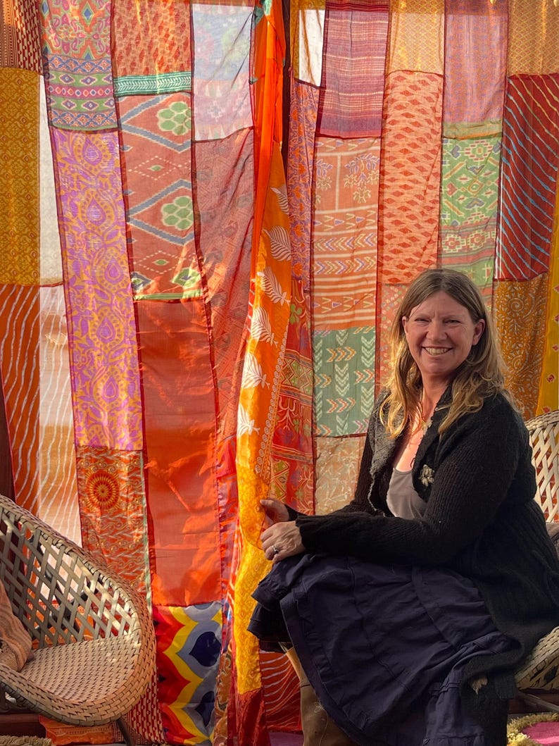 Pode incluir: Uma mulher est&aacute; sentada em uma cadeira de vime em frente a uma cortina de patchwork colorida. A cortina &eacute; feita de muitos tecidos diferentes em tons de laranja, vermelho, amarelo e roxo. A mulher est&aacute; vestindo um su&eacute;ter preto e uma saia longa e escura.