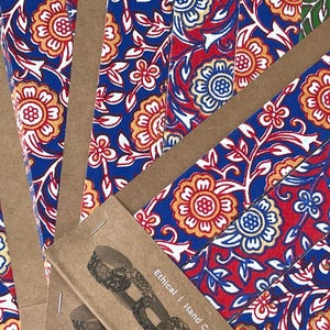 May include: Close-up of several notebooks with a vibrant floral pattern in red, blue, and gold. The notebooks are bound with brown paper and staples. The text "Ethical | Hand Crafted | Eco" is visible on the packaging.