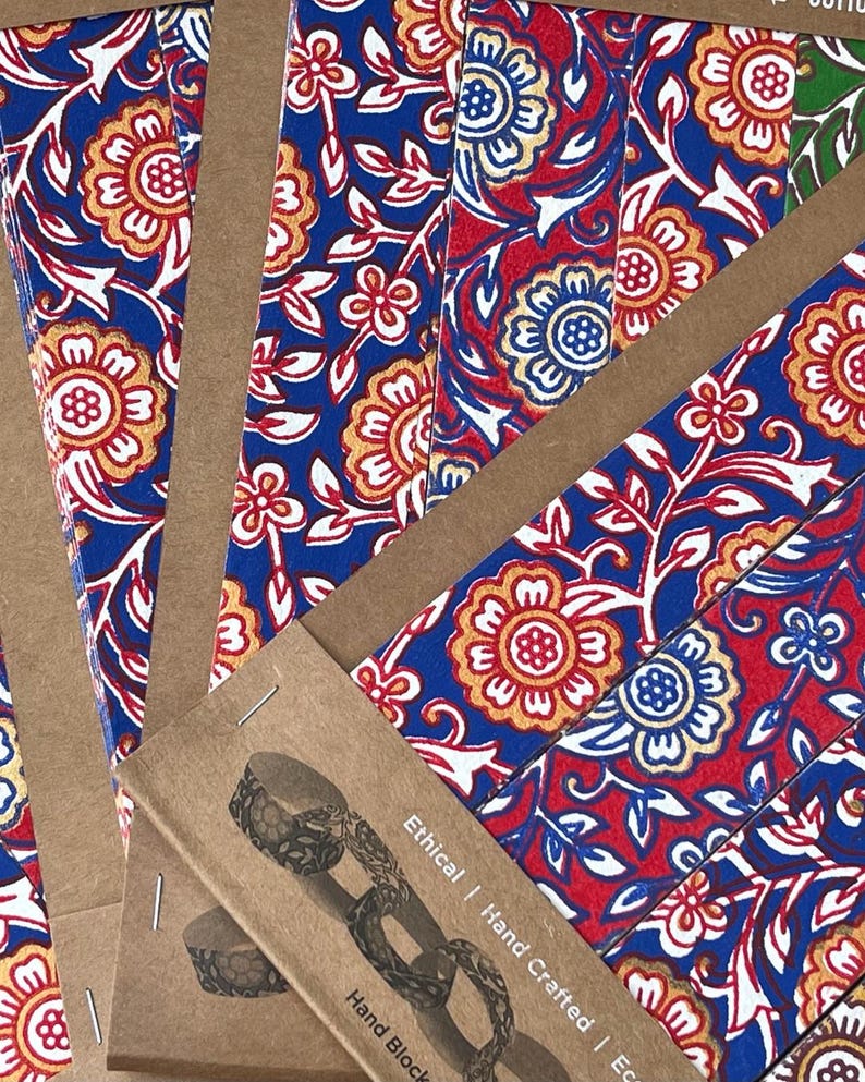 May include: Close-up of several notebooks with a vibrant floral pattern in red, blue, and gold. The notebooks are bound with brown paper and staples. The text "Ethical | Hand Crafted | Eco" is visible on the packaging.