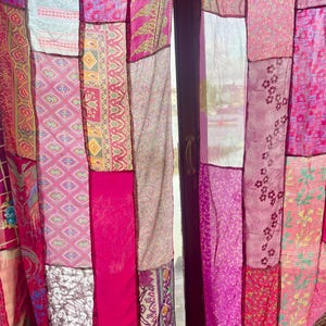 May include: A patchwork curtain made of various pink fabrics. The curtain is composed of rectangular panels with different patterns and shades of pink, creating a vibrant and textured appearance. The curtain hangs in front of a window, allowing light to filter through.