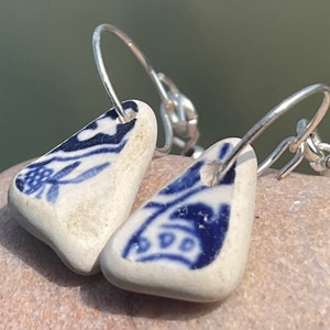 Puede incluir: Un par de pendientes de aro de plata con colgantes de cerámica azul y blanca. Los colgantes tienen forma de triángulo y presentan un diseño floral.