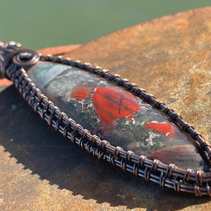 Puede incluir: Un colgante largo y ovalado con una piedra roja, negra y blanca engastada en un marco de alambre de cobre. El alambre está tejido en un patrón de cesta.