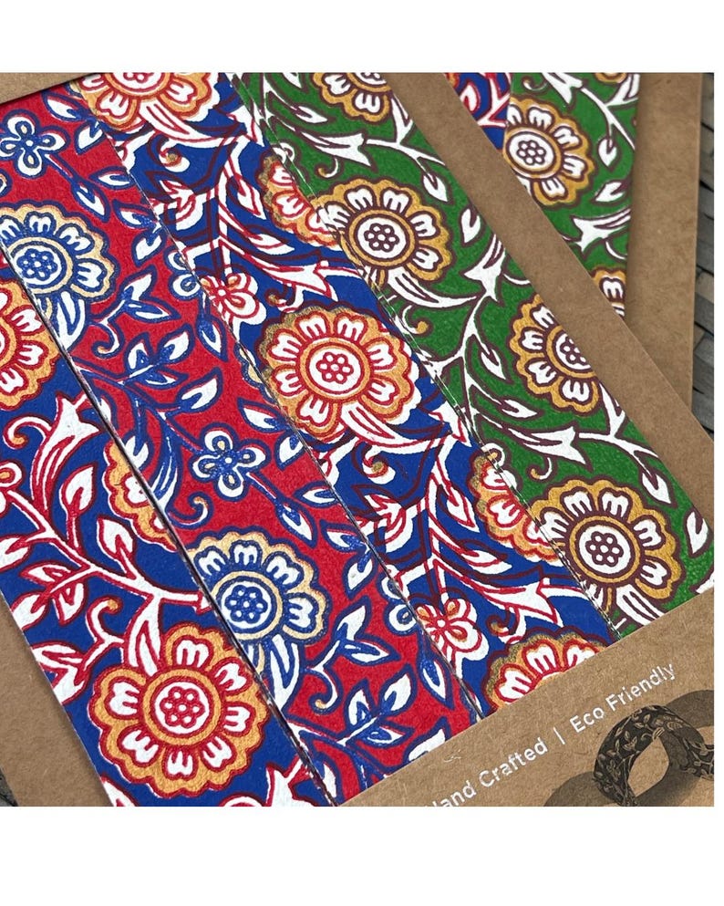 May include: Close-up of several floral patterned bookmarks. The bookmarks feature a repeating design of flowers and leaves in red, blue, gold, and green. The text "Hand Crafted | Eco Friendly" is visible.