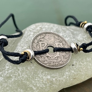 Puede incluir: Una pulsera de cordón negro con una moneda de plata como pieza central. La moneda tiene un diseño detallado con un agujero central y está acentuada con cuentas plateadas y doradas. La pulsera se muestra sobre una superficie verde claro.