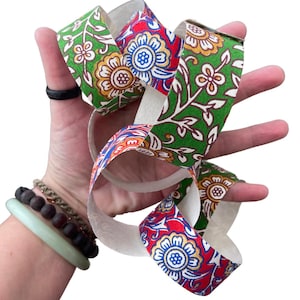 May include: A decorative ribbon with a floral pattern in green, red, yellow, and blue. The ribbon is held in a hand, showcasing its intricate design. The ribbon's design features a repeating pattern of flowers and leaves.