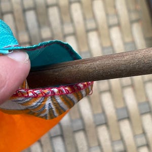 Pode incluir: Close-up de uma haste de madeira sendo inserida em um tubo de tecido colorido com costuras vermelhas. O tecido &eacute; laranja, branco e azul.