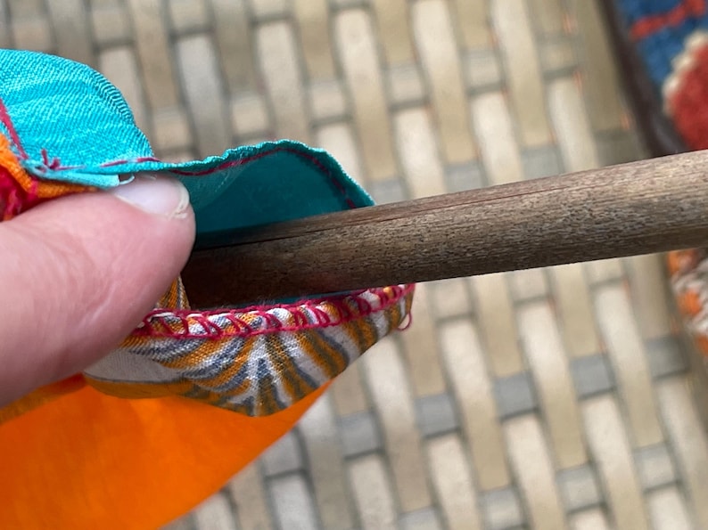 Pode incluir: Close-up de uma haste de madeira sendo inserida em um tubo de tecido colorido com costuras vermelhas. O tecido &eacute; laranja, branco e azul.