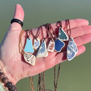 Puede incluir: Una colección de ocho collares con diferentes colgantes de cerámica. Los colgantes son de diferentes formas y tamaños, y están decorados con motivos florales azules y blancos. Los collares están hechos con cordones marrones.