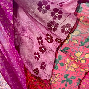 May include: Close-up of layered fabrics in shades of pink and purple. The fabrics feature floral patterns, including small white flowers and larger stylized blooms. The textures vary, with some appearing sheer and others more opaque.