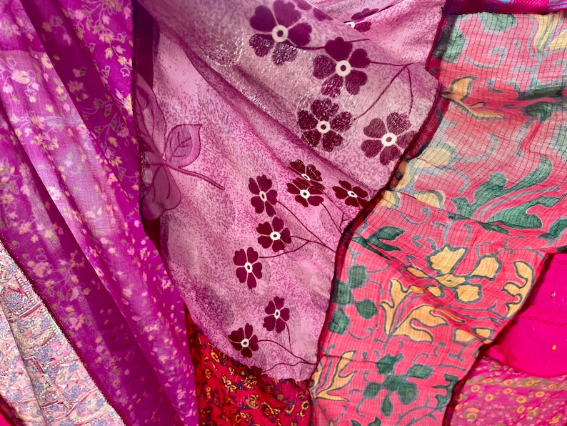 May include: Close-up of layered fabrics in shades of pink and purple. The fabrics feature floral patterns, including small white flowers and larger stylized blooms. The textures vary, with some appearing sheer and others more opaque.