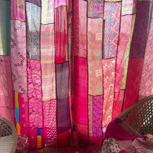 May include: A patchwork curtain made of various fabrics in shades of pink, magenta, and red. The curtain is composed of vertical panels with different patterns and textures, creating a vibrant and eclectic look. The overall theme is bohemian and decorative.