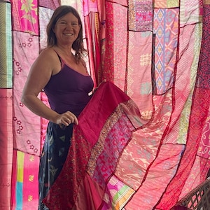 May include: A woman holding a vibrant, patchwork textile in shades of pink and red. The textile features various patterns and textures, with a sheer quality. The background is a curtain made of similar patchwork fabrics. The woman is wearing a purple tank top and a dark skirt.