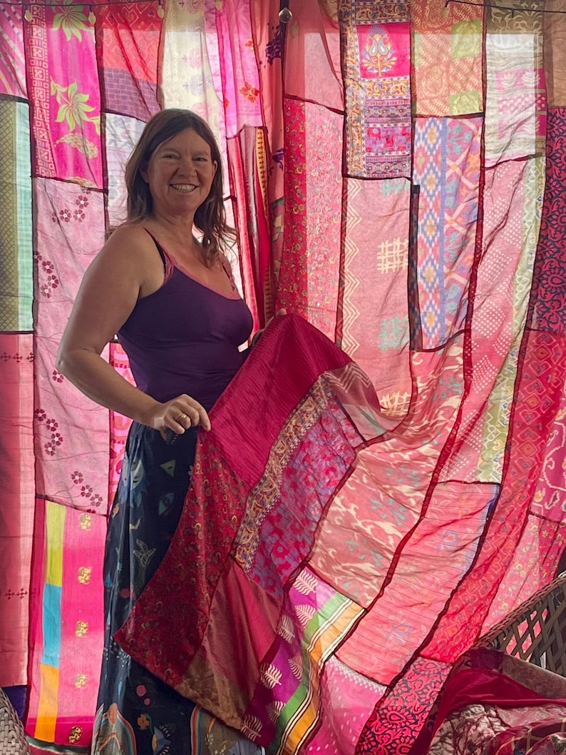 May include: A woman holding a vibrant, patchwork textile in shades of pink and red. The textile features various patterns and textures, with a sheer quality. The background is a curtain made of similar patchwork fabrics. The woman is wearing a purple tank top and a dark skirt.