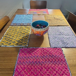 May include: A wooden table displays a collection of colorful placemats and a decorative bowl. The placemats feature various patterns and colors, including pink, yellow, blue, and purple. The bowl is painted with intricate designs and holds small decorative items.