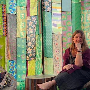 May include: A woman sits in front of a colourful patchwork curtain, her foot resting on a wooden stool. The curtain is made of various fabrics in shades of green, blue, and purple, with intricate patterns and designs.