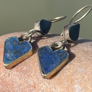 Könnte beinhalten: Silberne Ohrringe mit herzförmigen Anhängern aus Lapislazuli. Die Ohrringe haben einen dunkelblauen Stein, der in einen goldfarbenen Metallrahmen gefasst ist. Die Ohrringe werden auf einer strukturierten, hellbraunen Oberfläche präsentiert.