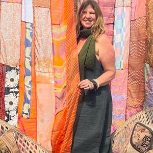 Pode incluir: Uma mulher vestindo um vestido verde escuro sem mangas e um len&ccedil;o est&aacute; em frente a uma cortina de patchwork colorida. Ela est&aacute; sorrindo e olhando para a c&acirc;mera. A cortina &eacute; feita de diferentes tecidos e cores, incluindo laranja, rosa e azul.