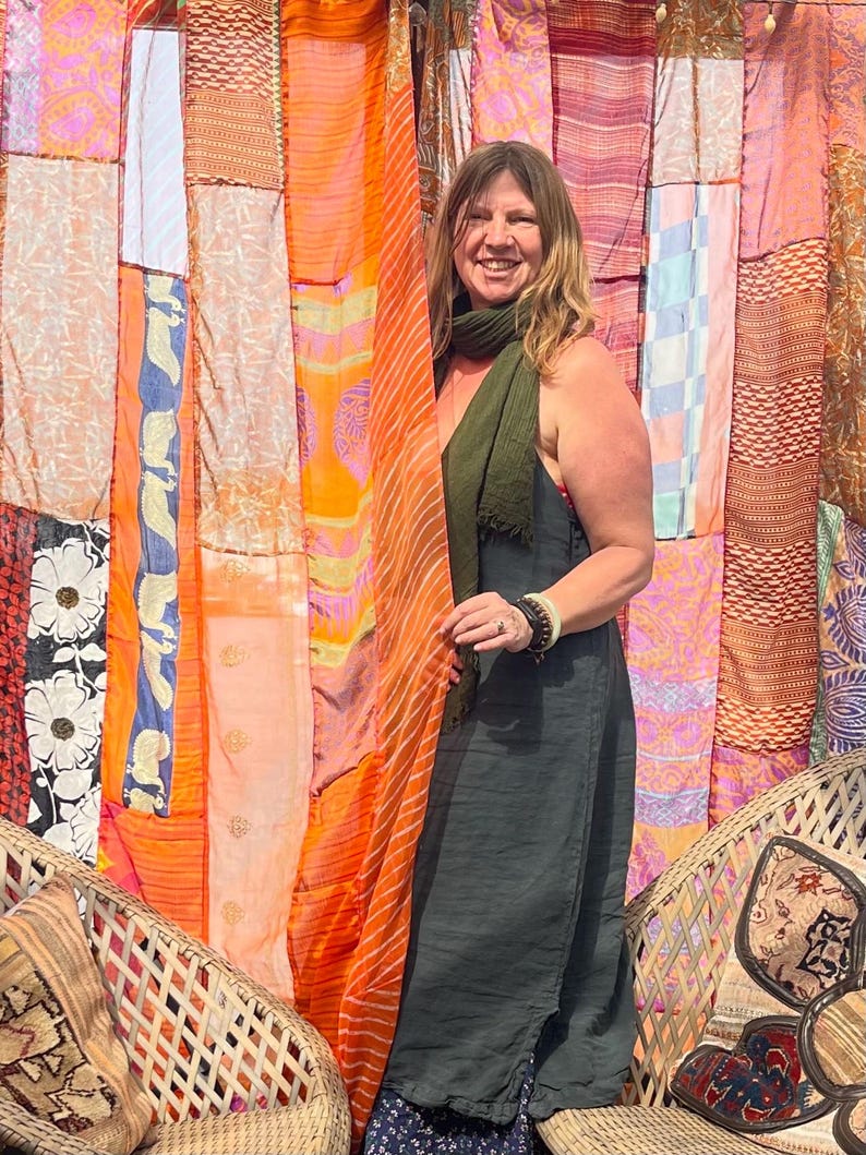 Pode incluir: Uma mulher vestindo um vestido verde escuro sem mangas e um len&ccedil;o est&aacute; em frente a uma cortina de patchwork colorida. Ela est&aacute; sorrindo e olhando para a c&acirc;mera. A cortina &eacute; feita de diferentes tecidos e cores, incluindo laranja, rosa e azul.