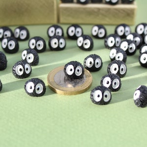 May include: A group of small, black, fuzzy, round figurines with white eyes. The figurines are scattered on a green surface with a euro coin in the center.