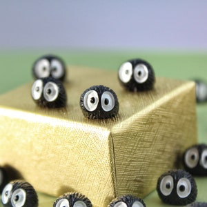 May include: A group of small, black, fuzzy, round ornaments with white eyes. The ornaments are arranged on a gold gift box and a green surface.