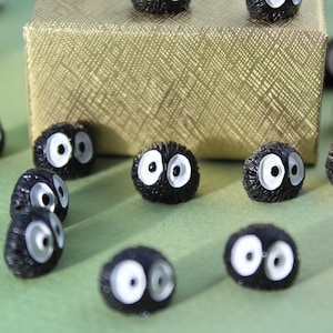 May include: A group of small, black, fuzzy, round ornaments with white eyes. The ornaments are scattered on a green surface and near a gold box.