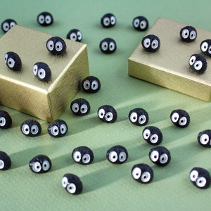 May include: A collection of small, black, fuzzy, round ornaments with white eyes. The ornaments are scattered on a green surface and some are on top of two gold gift boxes.